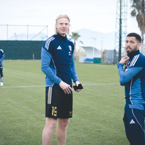 Dwaj piłkarze w strojach treningowych rozmawiają na boisku, podczas gdy w tle widać innych zawodników i otoczenie sportowe.