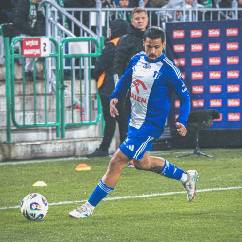 Na zdjęciu widać piłkarza w niebiesko-białym stroju, który wykonuje rzut rożny na boisku piłkarskim w deszczowej atmosferze.