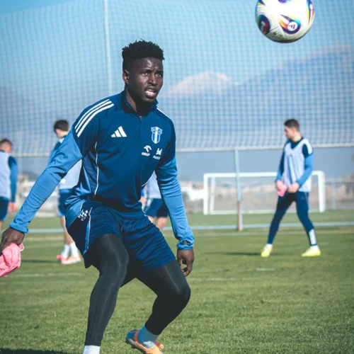 Na zdjęciu przedstawiony jest młody piłkarz podczas treningu na boisku, odbierający piłkę w jasnym otoczeniu górskim.