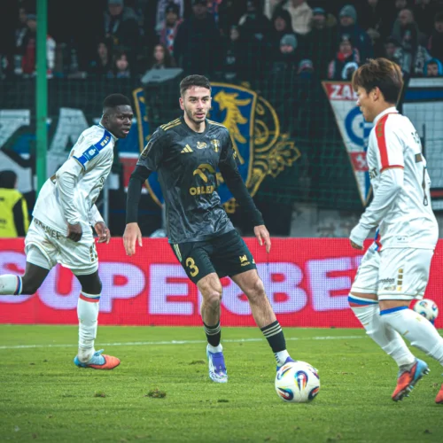 Trzech piłkarzy na boisku w trakcie meczu, z piłką w grze i kibicami w tle.