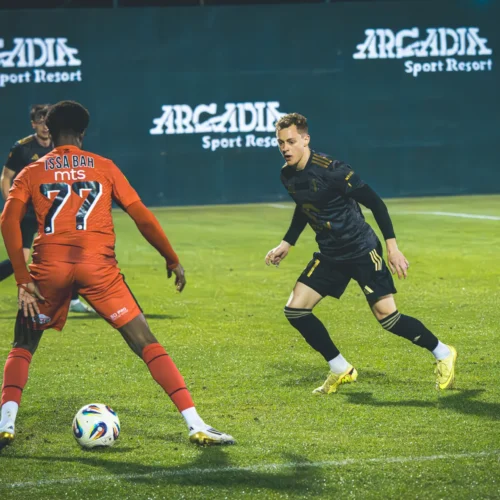 Na zdjęciu dwóch piłkarzy rywalizuje ze sobą na murawie, a w tle widoczny jest napis "Arcadia Sport Resort".