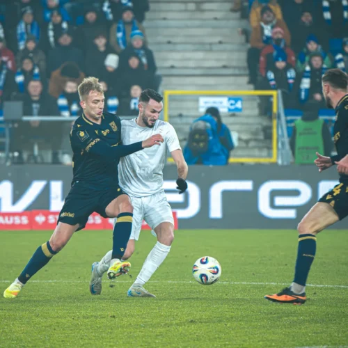 Na zdjęciu znajduje się scena z meczu piłkarskiego, na której zawodnicy walczą o piłkę, a w tle widać kibiców i stadion.