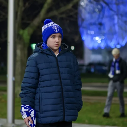 Młody chłopiec w niebieskiej czapce i puchowej kurtce stoi na tle oświetlonych drzew, trzymając w ręku niebieski szalik.