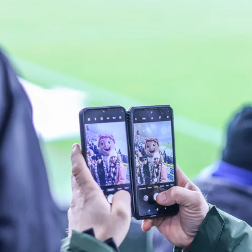 Dwie osoby robią zdjęcie z maskotką na stadionie, trzymając w rękach telefony.