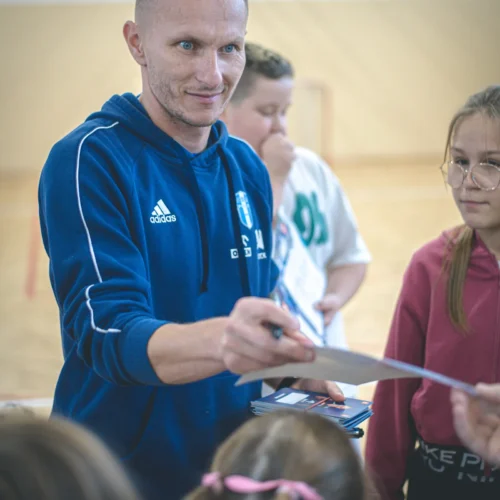 Na zdjęciu widoczny jest mężczyzna w niebieskiej bluzie Adidas, który z uśmiechem rozdaje materiały dzieciom w hali sportowej.
