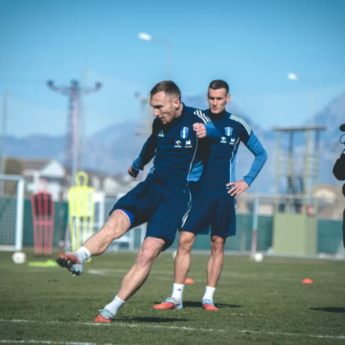 Dwaj piłkarze obserwują trenującego kolegę na boisku, podczas gdy jedna osoba rejestruje sytuację kamerą.