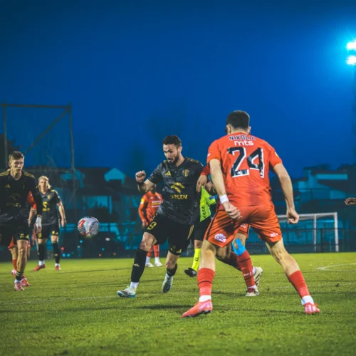 Na zdjęciu widać zawodników grających w piłkę nożną na boisku przy wieczornym oświetleniu.