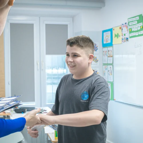 Chłopiec uśmiecha się, podając rękę postaci w kostiumie, w sali lekcyjnej pełnej kolorowych materiałów edukacyjnych.