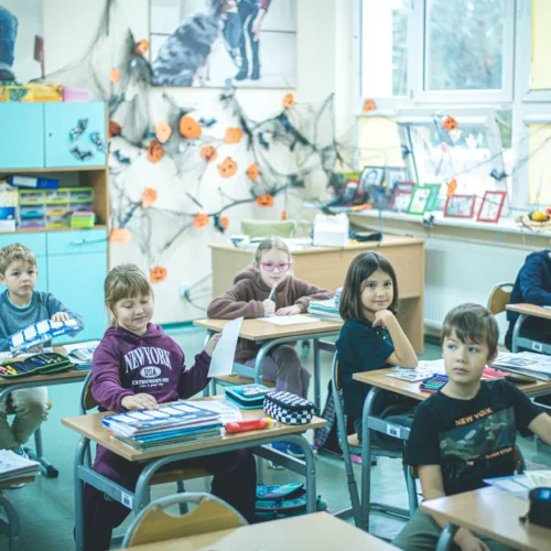 Na zdjęciu widać grupę dzieci siedzących przy biurkach w klasie, która jest udekorowana na Halloween.