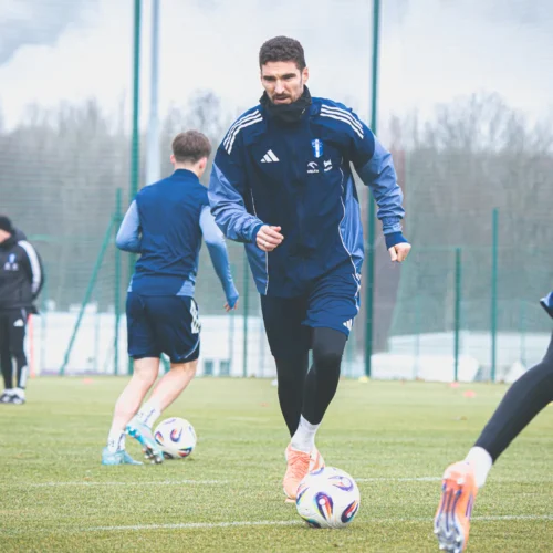 Na zdjęciu widać kilku piłkarzy trenujących na boisku, z jednym zawodnikiem biegnącym w kierunku piłki.