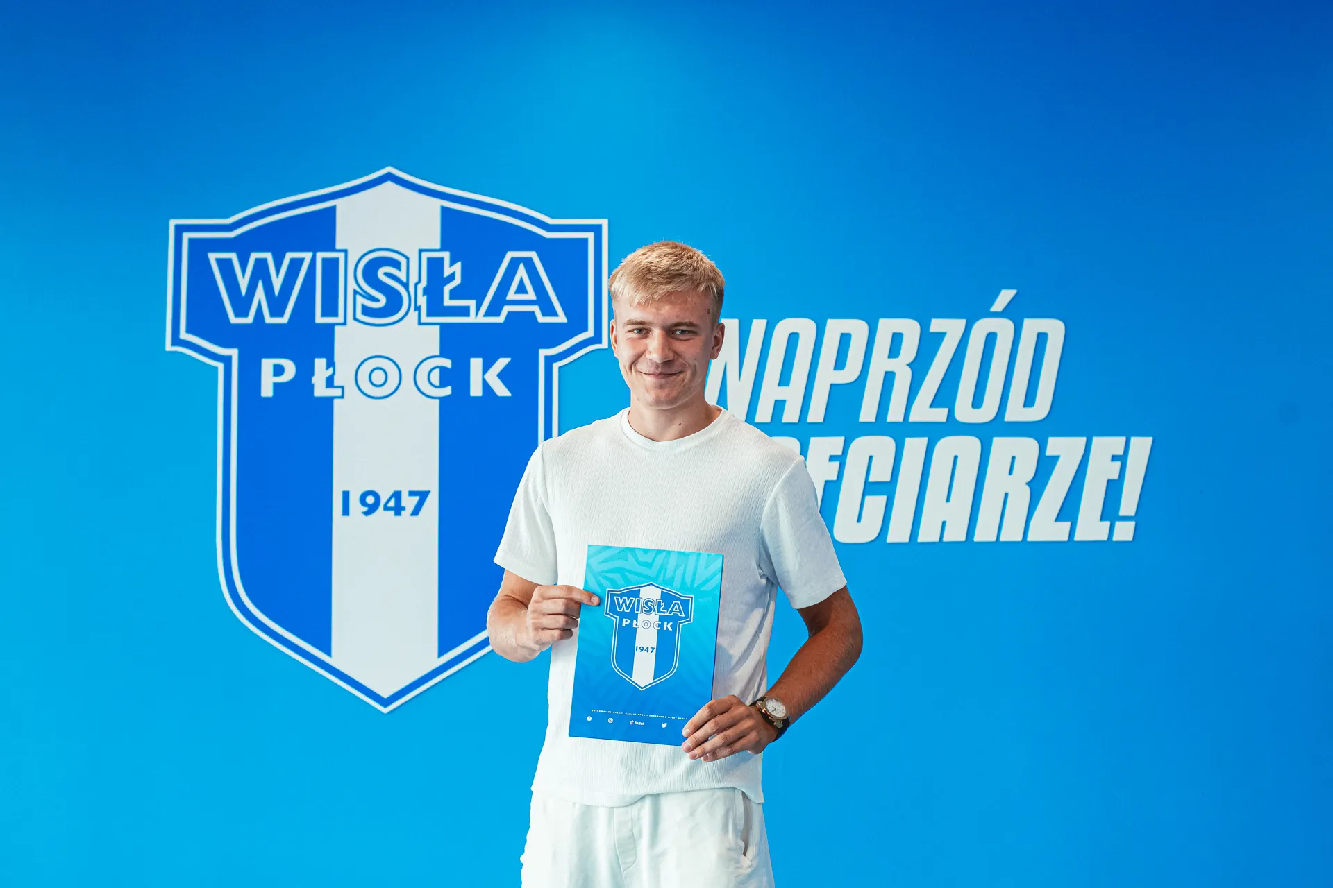 Na zdjęciu młody mężczyzna stoi z uśmiechem, trzymając dokument na tle niebieskiej ściany z logo klubu piłkarskiego Wisła Płock oraz hasłem „Naprzód, Płocczanie!”.