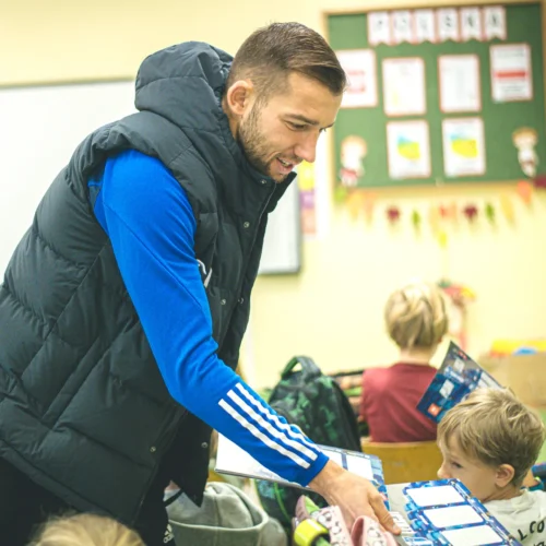Na zdjęciu mężczyzna w kamizelce rozmawia z dziećmi w klasie szkolnej.