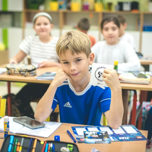 Chłopiec w niebieskiej koszulce uśmiecha się i pokazuje kartkę z logo, siedząc w klasie z kolegami w tle.