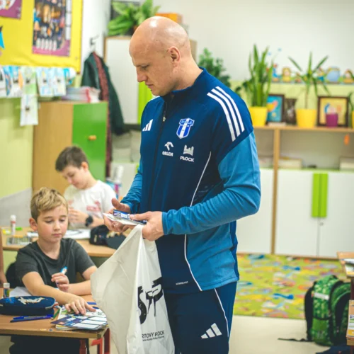 Na zdjęciu widoczny jest mężczyzna w niebieskim stroju sportowym, który z zainteresowaniem obserwuje dziecko pracujące w klasie na zajęciach plastycznych.