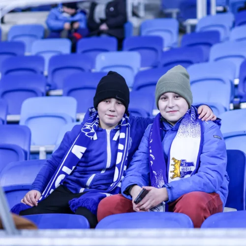 Dwoje dzieci w szalikach drużynowych siedzi na niebieskich siedzeniach stadionu, uśmiechając się do aparatu.