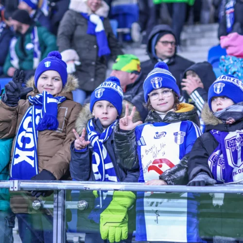 Na zdjęciu widać grupę radosnych dzieci w czapkach i szalikach kibiców, które celebrują mecz sportowy.