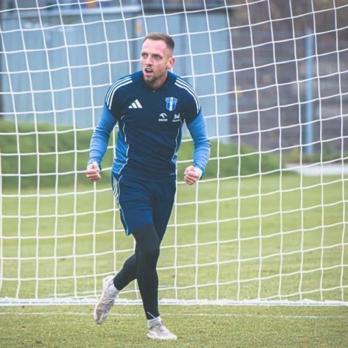 Na zdjęciu widać bramkarza w niebieskim stroju, który z entuzjazmem zaciąga się w kierunku bramki podczas treningu na boisku piłkarskim.