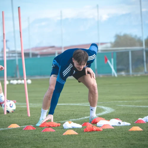 Dwaj młodzi sportowcy trenują na boisku piłkarskim, jednocześnie układając pachołki i przygotowując się do ćwiczeń.