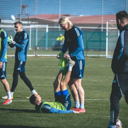 Na zdjęciu widać grupę piłkarzy podczas treningu na boisku, gdzie jeden z zawodników leży na ziemi, a inny pomaga mu w rozciąganiu.