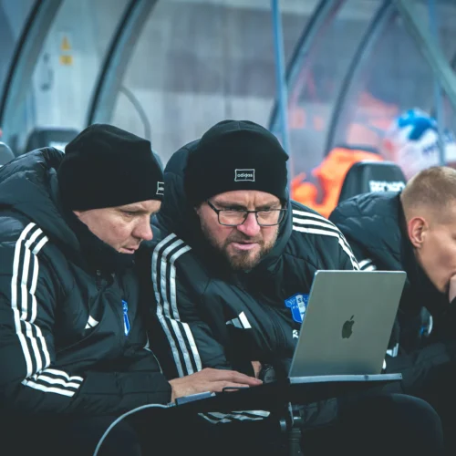 Na zdjęciu widać członków drużyny sportowej w strojach treningowych, którzy z uwagą analizują dane na laptopie, siedząc na ławce rezerwowych podczas meczu.