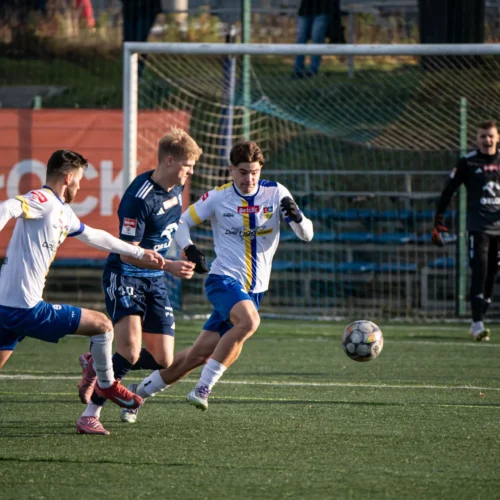 Na zdjęciu dwóch zawodników piłkarskich walczy o piłkę na boisku, a w tle widać bramkarza i resztę drużyny.
