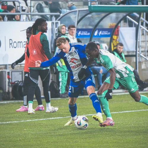 Na zdjęciu dwaj piłkarze zacięcie walczą o piłkę podczas meczu, w tle widać ławkę rezerwowych i trybuny pełne kibiców.