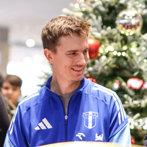 Na zdjęciu widać mężczyznę w niebieskiej bluzie, uśmiechającego się przy choince ozdobionej czerwonymi bombkami i światełkami.