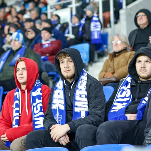 Grupa młodych kibiców w barwach swoich drużyn siedzi na trybunach, skoncentrowana na wydarzeniach na boisku.