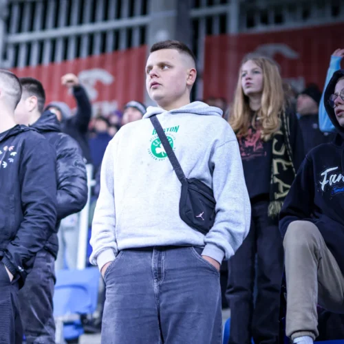 Na zdjęciu widać grupę kibiców na trybunach stadionu, a w centrum znajduje się młody mężczyzna w szarej bluzie z kapturem.