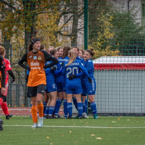 Na boisku piłkarskim, zawodniczki w niebieskich strojach świętują zdobycie bramki, podczas gdy bramkarka w pomarańczowym przygląda się z dezaprobatą.