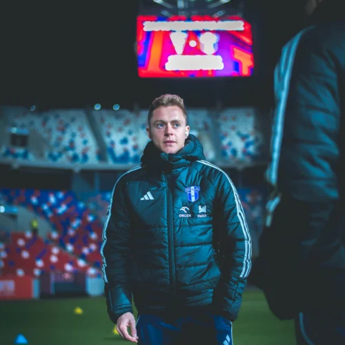 Mężczyzna w sportowej kurtce stoi na boisku piłkarskim, a w tle widoczna jest wielka tablica elektroniczna.