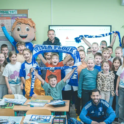 Na zdjęciu widać grupę uśmiechniętych dzieci w klasie, które trzymają szaliki drużyny Wisła Płock, a wśród nich znajdują się także dwaj mężczyźni oraz maskotka drużyny.