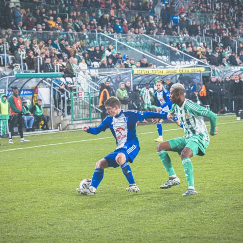 Na zdjęciu widać dwóch piłkarzy podczas meczu, jeden z nich w niebieskim, a drugi w zielonym stroju, starających się przejąć piłkę na tle wypełnionego kibicami stadionu.