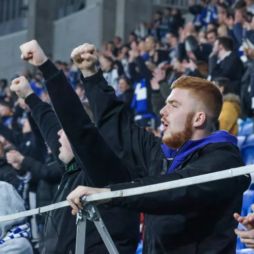 Grupa kibiców na stadionie entuzjastycznie podnosi ręce w górę, celebrując emocje związane z meczem.