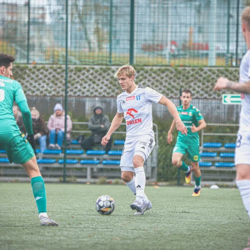 Na zdjęciu widać zawodników grających w piłkę nożną na boisku, z jednym graczem w białym stroju w akcji z piłką, a w tle znajdują się zawodnicy w zielonych strojach oraz kilku widzów.