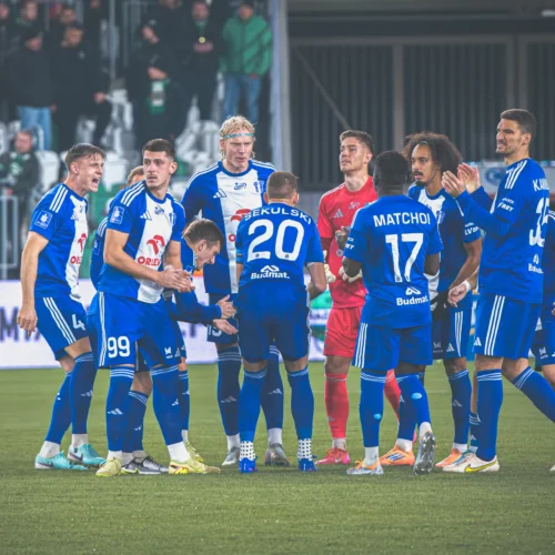 Grupa piłkarzy w niebieskich strojach celebruje zdobycie gola na boisku piłkarskim podczas meczu.