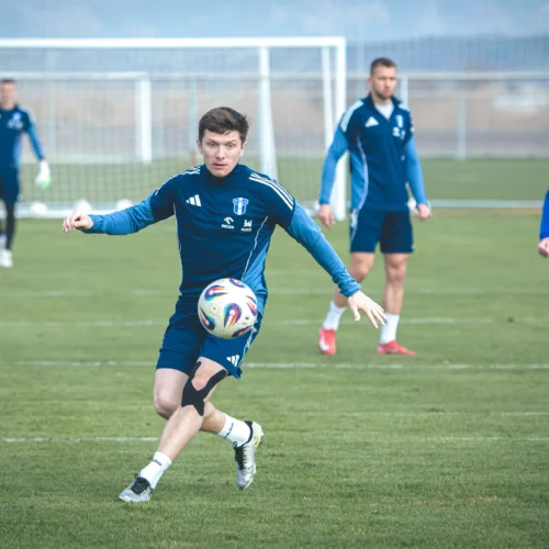 Na zdjęciu widać piłkarza wykonującego podanie podczas treningu na boisku, z innymi zawodnikami w tle.