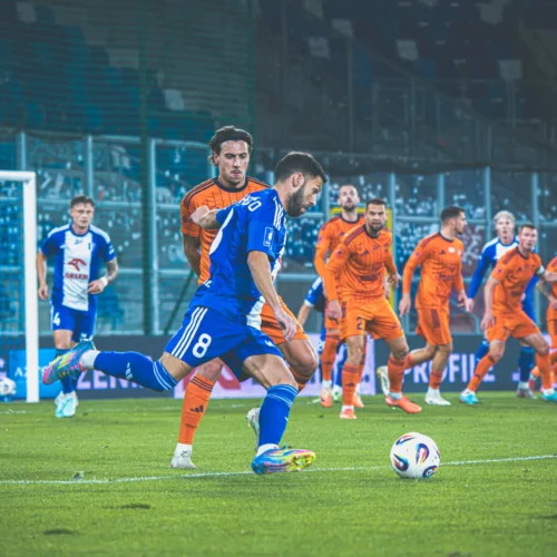 Na zdjęciu widoczna jest akcja podczas meczu piłkarskiego, z zawodnikami w niebieskich i pomarańczowych strojach, a także piłką leżącą na trawie.