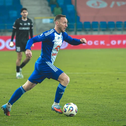 Na zdjęciu piłkarz w niebieskim stroju biegnie z piłką na boisku, podczas gdy bramkarz w czarnym stroju obserwuje sytuację.