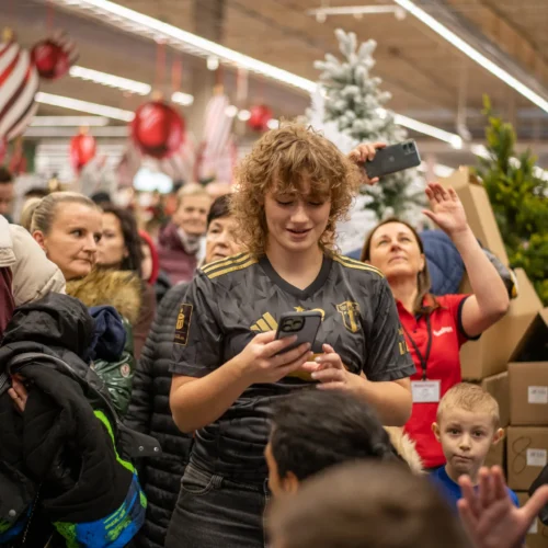 Zdjęcie przedstawia tłum ludzi w sklepie, z wyraźnym skupieniem na młodej osobie w czarnej koszulce, trzymającej telefon i uśmiechającej się wśród zabawek i dekoracji świątecznych.