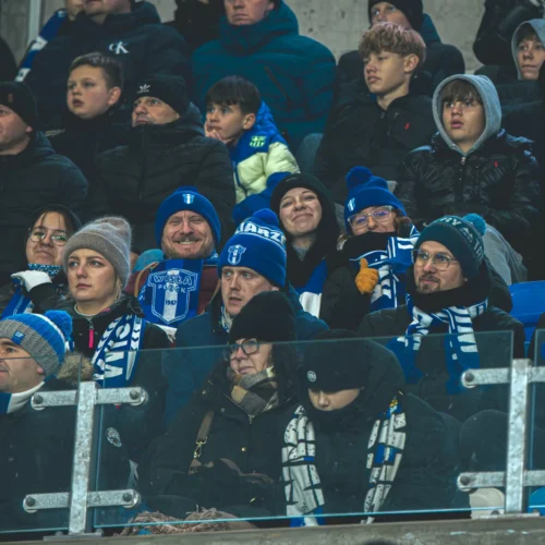 Na zdjęciu widać grupę kibiców ubranych w szaliki i czapki, którzy oglądają mecz na stadionie.