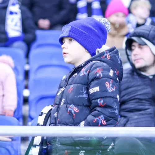 Na zdjęciu widoczny jest chłopiec w niebieskiej czapce i kurtce z motywem Marvela, który ogląda wydarzenie sportowe wśród zasiadających na trybunach widzów.