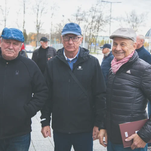 Trzech mężczyzn w ciepłych ubraniach stoi na zewnątrz, uśmiechając się, w tle widać innych mężczyzn i miejskie otoczenie.