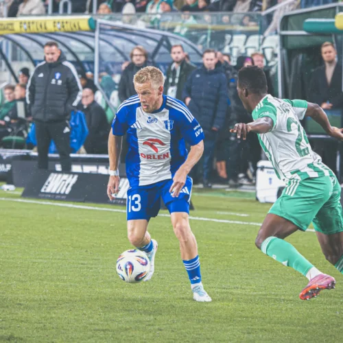 Na zdjęciu widać dwóch piłkarzy podczas meczu, jeden z nich w niebiesko-białej koszulce, a drugi w zielono-białej, w momencie, gdy jeden z graczy próbuje przejąć piłkę.