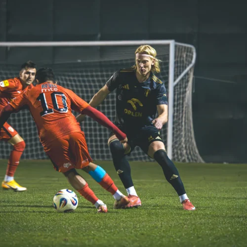 Na zdjęciu widać dwóch piłkarzy w trakcie dynamicznej akcji na boisku, z jednym z zawodników w czerwonym stroju próbującym ominąć przeciwnika w czarnym uniformie.