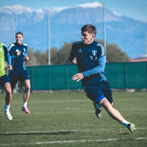 Na zdjęciu trzej piłkarze w granatowych strojach biegają po boisku treningowym, w tle widać góry pokryte śniegiem.