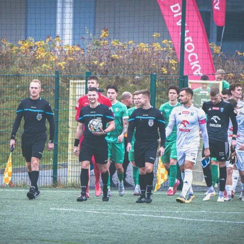 Na zdjęciu widoczna jest drużyna piłkarska oraz sędziowie wchodzący na boisko w otoczeniu flag i niesprzyjającej pogody.