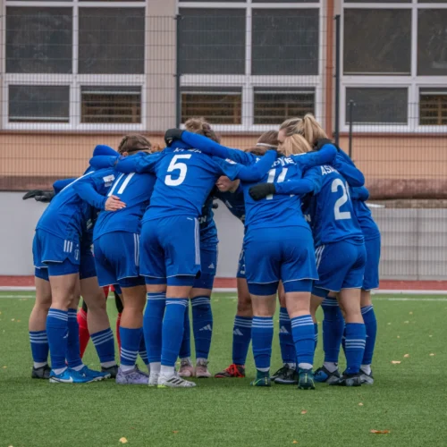 Grupa zawodniczek w niebieskich strojach sportowych tworzy krąg, przygotowując się do meczu na boisku ze sztuczną nawierzchnią.