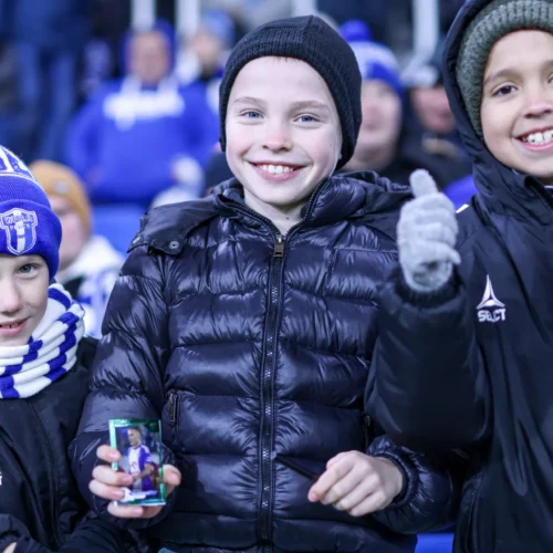 Na zdjęciu trzech uśmiechniętych chłopców stoi na trybunie, trzymając kartkę i pokazując gest „thumbs up” podczas meczu.