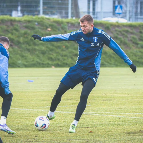 Na zdjęciu widać dwóch piłkarzy podczas treningu na boisku, z jednym z nich wykonującym podanie.